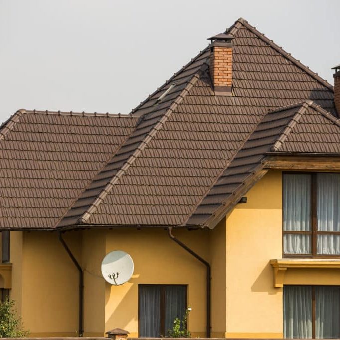 Top of big modern expensive residential house with shingled brown roof, high brick chimneys, stucco walls and plastic attic windows on blue sky copy space background. Real estate selling concept.